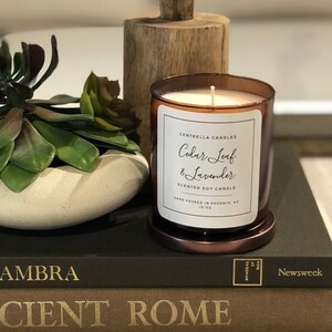 May include: A brown glass candle jar with a white label that reads "Centrella Candles Cedar Leaf & Lavender Scented Soy Candle Hand Poured in Phoenix, AZ 10 oz". The candle is sitting on a stack of books with a green succulent plant in a white pot on top.