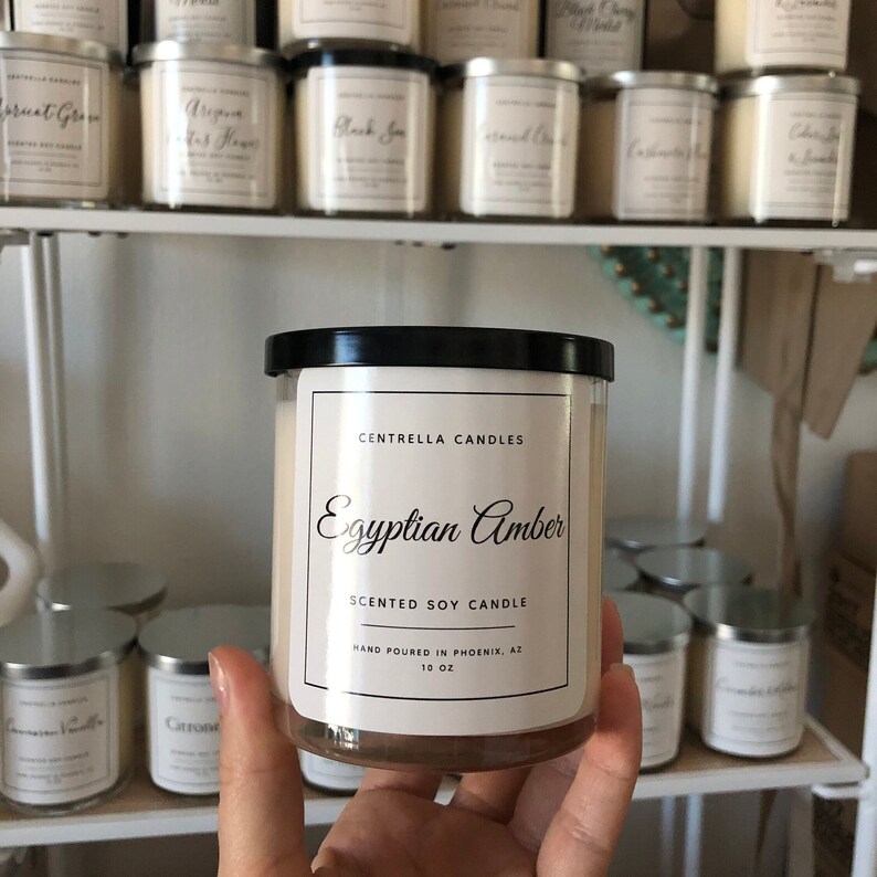 May include: A clear glass jar candle with a black lid, labeled "Egyptian Amber" by Centrella Candles. The label also states "Scented Soy Candle" and "10 OZ". Other candles are visible on a shelf in the background.