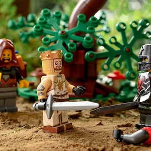 May include: A Lego scene featuring three minifigures: a king with a crown and sword, a hooded figure, and a black knight. The knight has a broken arm. The background includes a tree and other Lego elements.