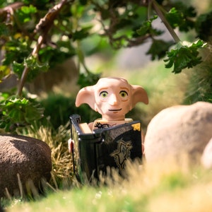 May include: A small, light-skinned elf figurine with large ears and green eyes is reading a black book with gold accents. The elf is sitting in a miniature forest setting with green plants and rocks.