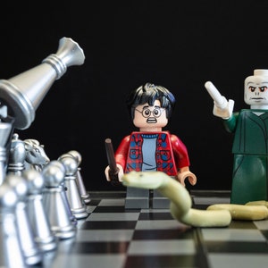 May include: A Lego Harry Potter minifigure with a wand and a snake, facing a Lego Voldemort minifigure with a wand. The minifigures are on a chessboard with silver and gold chess pieces.