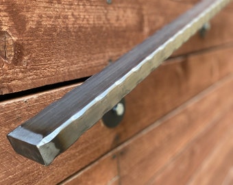 May include: A close-up of a metal handrail mounted to a wooden wall. The handrail is made of a thick, textured metal and has a square shape. The handrail is attached to the wall with a black metal bracket.