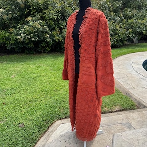 May include: A long, open-front rust-colored cardigan with a textured, patterned design. The cardigan has long sleeves and fringe detailing along the edges and hem. The garment is displayed on a black mannequin in an outdoor setting.