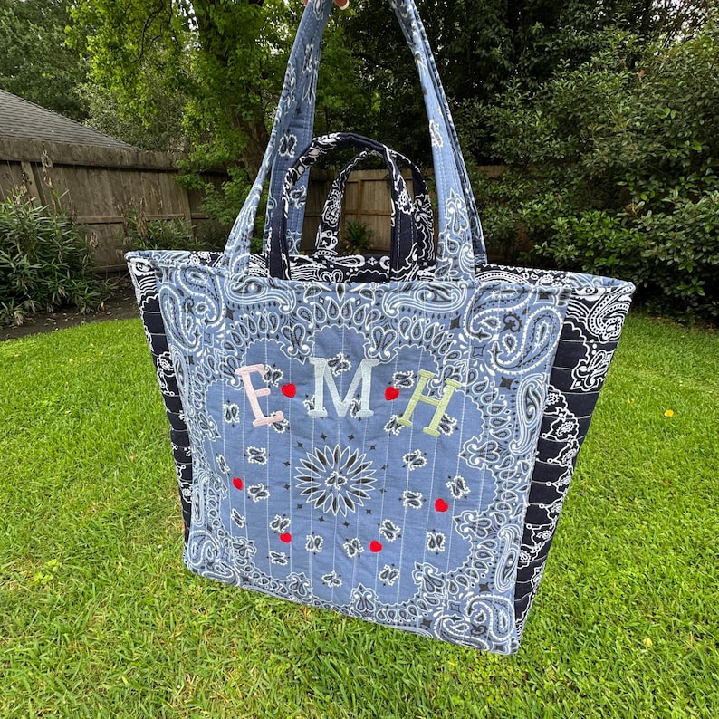 Puede incluir: Un bolso tote acolchado grande con un estampado de cachemira azul claro y las letras bordadas "EMH". El bolso tiene un bolso m&aacute;s peque&ntilde;o a juego en el interior y un estampado de cachemira negro en los laterales. El bolso tiene correas largas.