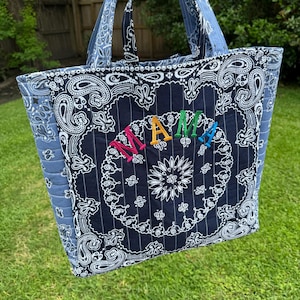 May include: A quilted tote bag with a blue paisley pattern and the word "MAMA" embroidered in rainbow colors. The bag has matching blue handles and a floral design in the center.