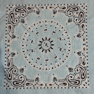Puede incluir: Un pa&ntilde;uelo cuadrado azul claro con un detallado estampado de cachemira en blanco y negro. El dise&ntilde;o presenta un motivo floral central y elementos de borde ornamentados, creando un textil decorativo cl&aacute;sico.