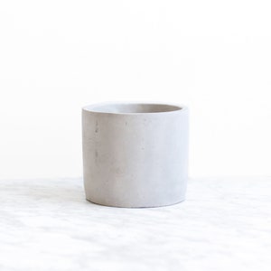 May include: A light gray concrete planter with a cylindrical shape. The planter is smooth and has a slightly textured surface.
