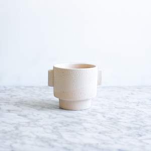 May include: A white ceramic planter with a textured finish and two handles on the sides. The planter has a square base and a round top.