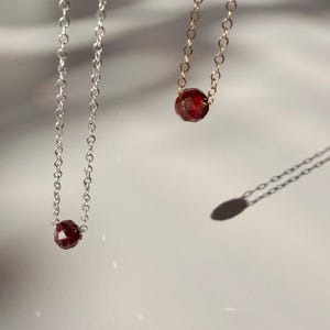 May include: Two delicate necklaces with faceted, deep red gemstones. One necklace features a silver chain, while the other has a gold chain. The gemstones are the focal point, suspended at the center of each chain.