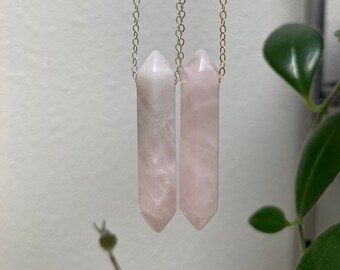 May include: Two pink crystal point pendants on silver and gold chains. The pendants are long and narrow, with a pointed tip.