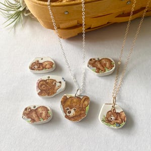 May include: Three necklaces with ceramic bear pendants. The pendants feature different sleeping bear illustrations. The necklaces are on a white surface with a wicker basket in the background.