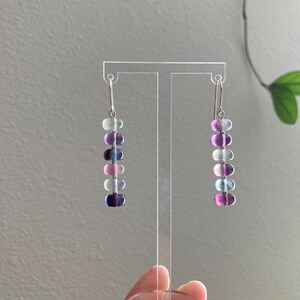 Op de afbeelding: Een paar oorhangers met een zilveren haak en een aflopende rij van zes ovale kralen in tinten paars, blauw en wit.