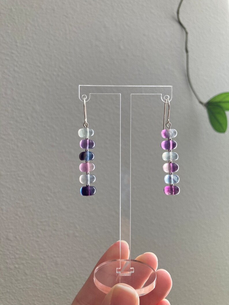 Op de afbeelding: Een paar oorhangers met een zilveren haak en een aflopende rij van zes ovale kralen in tinten paars, blauw en wit.