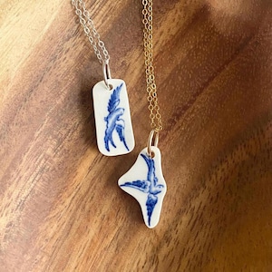 Könnte beinhalten: Zwei Halsketten mit weißen Anhängern mit blauen Vogelmotiven. Eine Kette hat eine silberne Kette, die andere eine goldene Kette. Die Anhänger haben eine unregelmäßige Form und ähneln zerbrochenen Porzellanstücken.