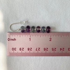 Op de afbeelding: Een paar oorbellen met een zilveren haak en een rij van zes paarse, grijze en transparante kralen. De oorbellen zijn ongeveer 2,5 cm lang.