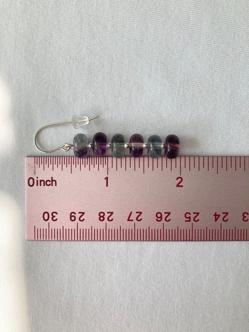 Op de afbeelding: Een paar oorbellen met een zilveren haak en een rij van zes paarse, grijze en transparante kralen. De oorbellen zijn ongeveer 2,5 cm lang.