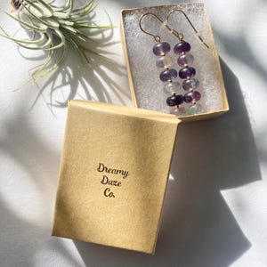 Op de afbeelding: Een paar lange, hangende oorbellen met een gradi&euml;nt van paarse, blauwe en groene fluorite kralen. De oorbellen zitten in een klein, bruin doosje met de tekst "Dreamy Daze Co." erop gedrukt.