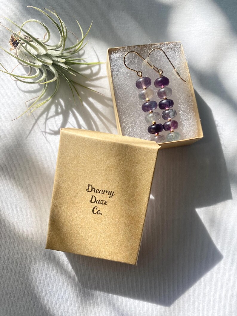 Op de afbeelding: Een paar lange, hangende oorbellen met een gradi&euml;nt van paarse, blauwe en groene fluorite kralen. De oorbellen zitten in een klein, bruin doosje met de tekst "Dreamy Daze Co." erop gedrukt.