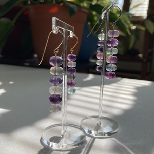 Op de afbeelding: Een paar hangende oorbellen met een gradi&euml;nt van paarse, blauwe en heldere edelstenen. De oorbellen worden getoond op een heldere acrylstandaard.
