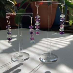 Op de afbeelding: Drie paar oorhangers met aflopende kralen. De kralen zijn iriserend en vari&euml;ren in kleur van lichtgroen tot paars. De oorhangers worden getoond op een transparante acrylstandaard.