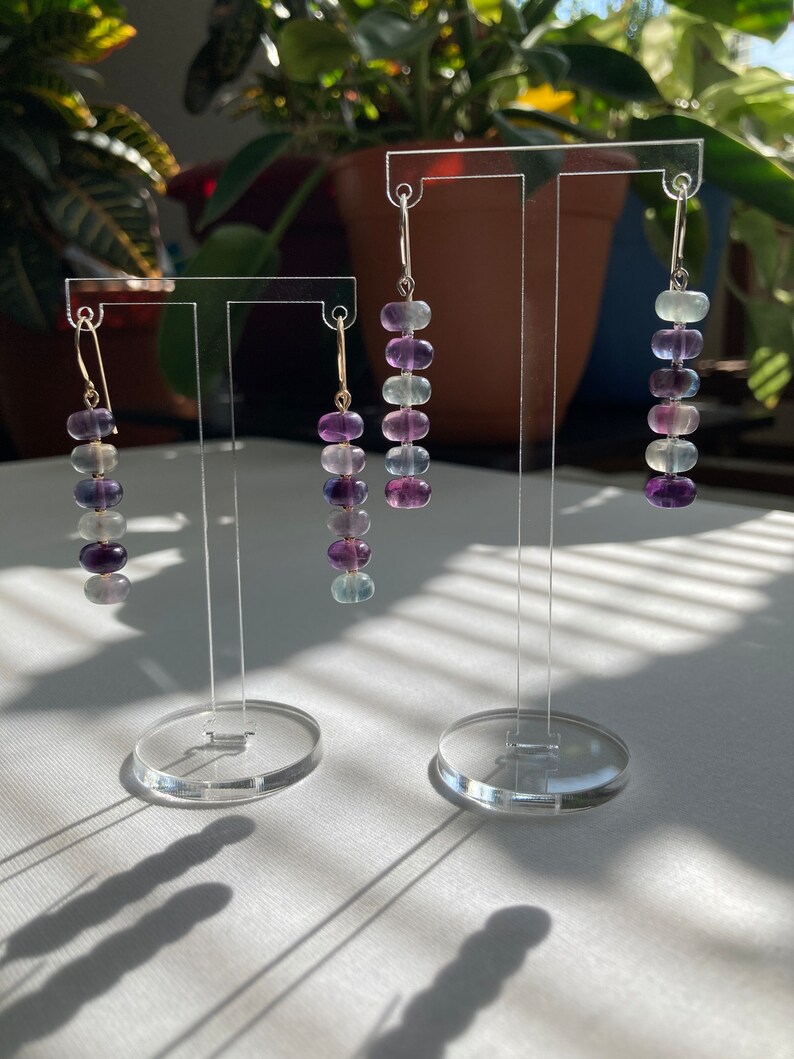 Op de afbeelding: Drie paar oorhangers met aflopende kralen. De kralen zijn iriserend en vari&euml;ren in kleur van lichtgroen tot paars. De oorhangers worden getoond op een transparante acrylstandaard.