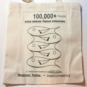 May include: A white canvas tote bag with black text and a graphic of stacked fish. The text reads "100,000+ People Need Organ/Tissue Donation". The bottom of the bag has the text "Image is the property of impathic images. Register Today - ImpathicImages.org"