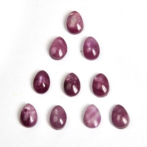 Op de afbeelding: Tien paarse edelsteen cabochons in een traanvorm, gerangschikt in een driehoekige vorm op een witte achtergrond.