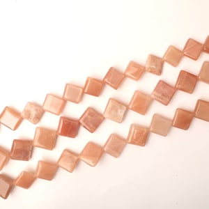 Puede incluir: Una hebra de cuentas de piedra de sol de color melocotón, cada cuenta es un cuadrado con bordes redondeados y forma de diamante.