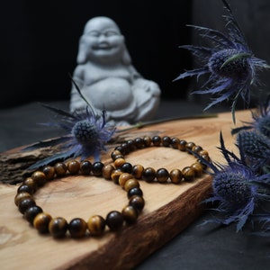 May include: Two brown and gold tiger's eye beaded bracelets on a wooden surface with a gray stone Buddha statue and blue thistle flowers in the background.
