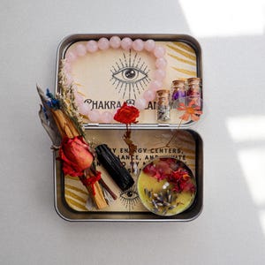 May include: A metal tin box filled with various items for a chakra balancing ritual. The box contains a pink rose quartz bracelet, a small candle, a bundle of dried herbs, a black obsidian crystal, and several small glass vials filled with dried flowers. The lid of the box features a graphic of an eye with the words "Chakra Balance" printed below.