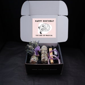 May include: A black gift box with a white card that says "Happy Birthday You Are So Magical" with a black and white illustration of a cat. The box contains a bundle of dried white sage, a bundle of dried lavender, a wooden stick of incense, a pink rose quartz bracelet, and an amethyst bracelet.