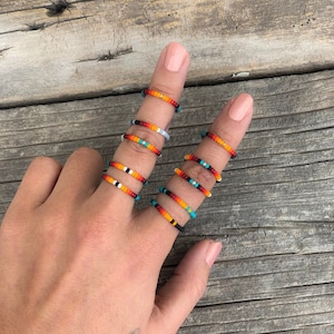 May include: A hand wearing a stack of colourful beaded rings. The rings are made of small, round beads in shades of orange, red, black, white, and turquoise. The rings are stacked on the middle finger of the hand.