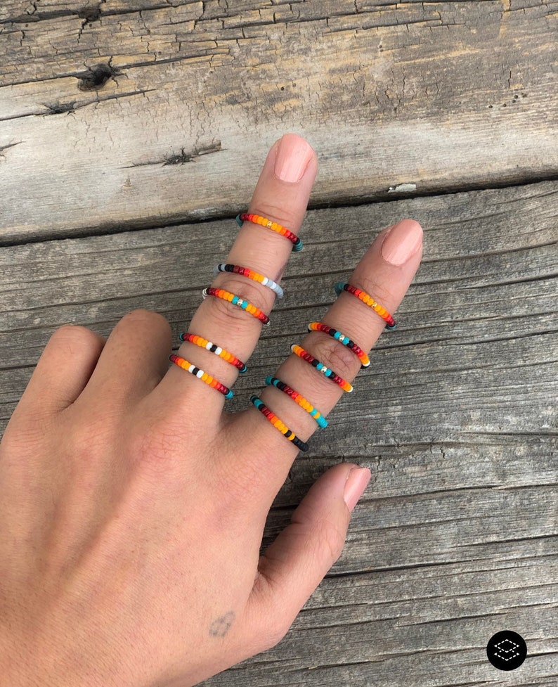 May include: A hand wearing a stack of colourful beaded rings. The rings are made of small, round beads in shades of orange, red, black, white, and turquoise. The rings are stacked on the middle finger of the hand.