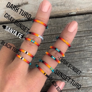 May include: A hand wearing six beaded rings with different colour combinations. The rings are labelled "Dark Turq", "Gray Sunset", "Santa Fe", "Black Turq", "Dusk Sunset", and "Sunset Turq".