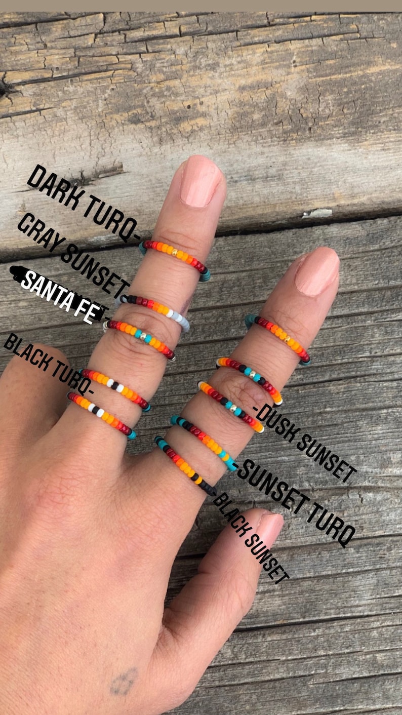 May include: A hand wearing six beaded rings with different colour combinations. The rings are labelled "Dark Turq", "Gray Sunset", "Santa Fe", "Black Turq", "Dusk Sunset", and "Sunset Turq".