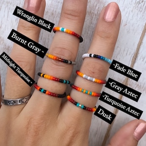 May include: A hand model wearing six different beaded rings. The rings are labeled with names like "Wranglin Black", "Burnt Gray", "Midnight Turquoise", "Fade Blue", "Gray Aztec", and "Turquoise Aztec".