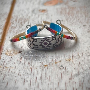 Puede incluir: Un anillo de plata con un patrón geométrico y una piedra turquesa. El anillo está rodeado por dos pulseras de cuentas, una blanca y otra con cuentas rojas, naranjas y turquesas.