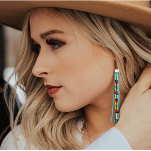 Könnte beinhalten: Eine Frau trägt einen braunen Hut und ein weißes Oberteil mit langen Perlenohrringen. Die Ohrringe sind türkis, rot, orange, gelb, weiß und schwarz.