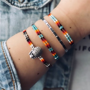 May include: A stack of colourful beaded bracelets with a silver charm in the shape of a Native American headdress. The bracelets are layered on a person's wrist.