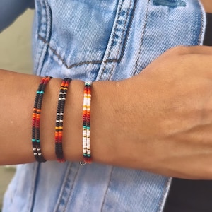 May include: Three beaded bracelets in a variety of colors, including black, red, orange, white, and turquoise. The bracelets are worn on a wrist against a denim jacket background. The bracelets are made of small, colorful beads.