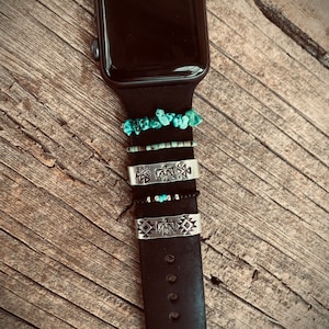 Può includere: Un Apple Watch nero con un cinturino nero. Il cinturino ha diversi anelli turchesi e argentati con disegni geometrici.