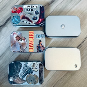 May include: Four small, silver, rectangular tins with different designs. The top tin has a red, white, and blue design with the words "Cowboy Bar". The second tin has a woman in a white fur coat with the word "Cowgirl" on the side. The third tin is plain silver. The fourth tin has a brown boot, a gun, and the words "Deadwood" on the side.