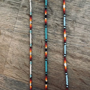 Puede incluir: Tres collares de cuentas con diferentes patrones de colores. Los collares están hechos con cuentas blancas, negras, rojas, naranjas y azules. Los collares están colgados sobre un fondo de madera.