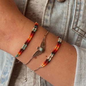 May include: Three bracelets on a wrist. The top bracelet is a multi-coloured beaded bracelet with red, orange, yellow, blue, and black beads. The middle bracelet is a silver chain with a silver eagle charm. The bottom bracelet is a multi-coloured beaded bracelet with red, orange, yellow, and white beads.
