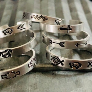 May include: A set of six silver rings with different Native American inspired designs. The designs include a buffalo, an arrow, a cross, and a geometric pattern.