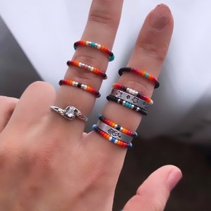 May include: A hand displays a collection of beaded rings in various colors, including red, orange, turquoise, and black. Some rings feature silver bands with star and arrow designs. A silver engagement ring is also visible.