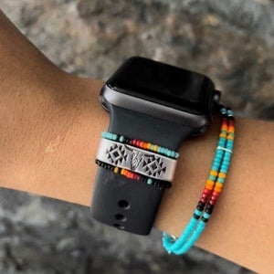 Puede incluir: Reloj inteligente negro con una banda plateada con un diseño geométrico y una calavera de toro. Una pulsera de cuentas en turquesa, naranja, rojo y negro está unida al reloj. El reloj está en una muñeca.
