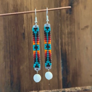 Pode incluir: Um par de brincos de contas com um padrão geométrico turquesa, vermelho, laranja e preto. Os brincos têm um gancho prateado e uma conta branca na parte inferior.