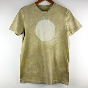 May include: A short-sleeved, olive green t-shirt with a large, off-white circle design on the front. The shirt is hanging on a wooden hanger. The fabric has a slightly faded appearance.