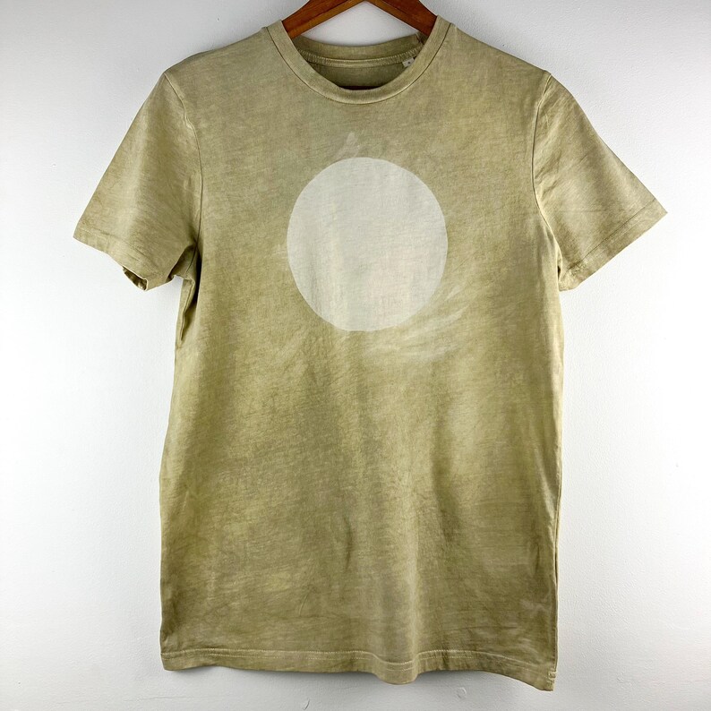 May include: A short-sleeved, olive green t-shirt with a large, off-white circle design on the front. The shirt is hanging on a wooden hanger. The fabric has a slightly faded appearance.
